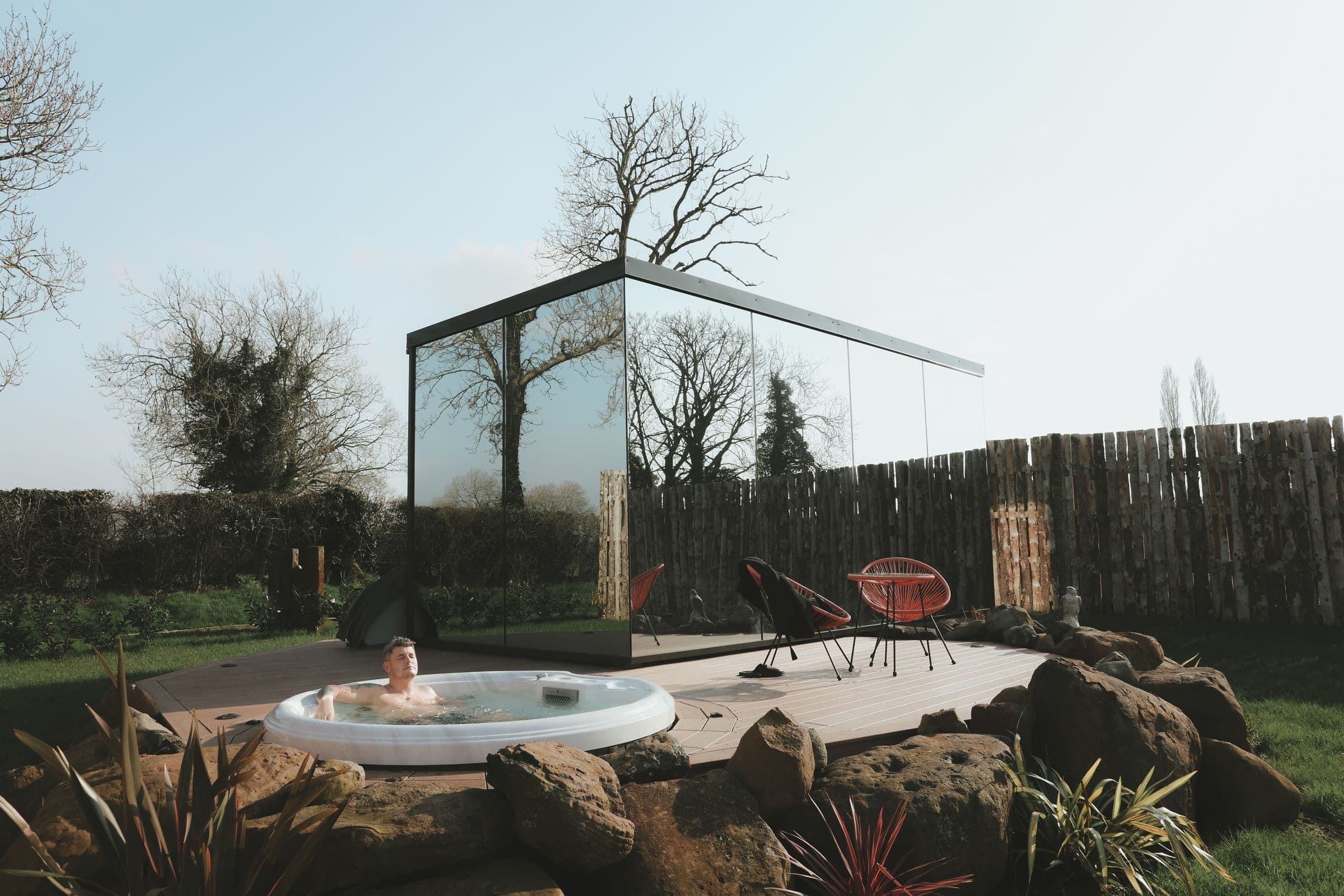The Sunrise mirror house reflecting the bare trees around it, with the round hot tub sunk into the timber decking in front