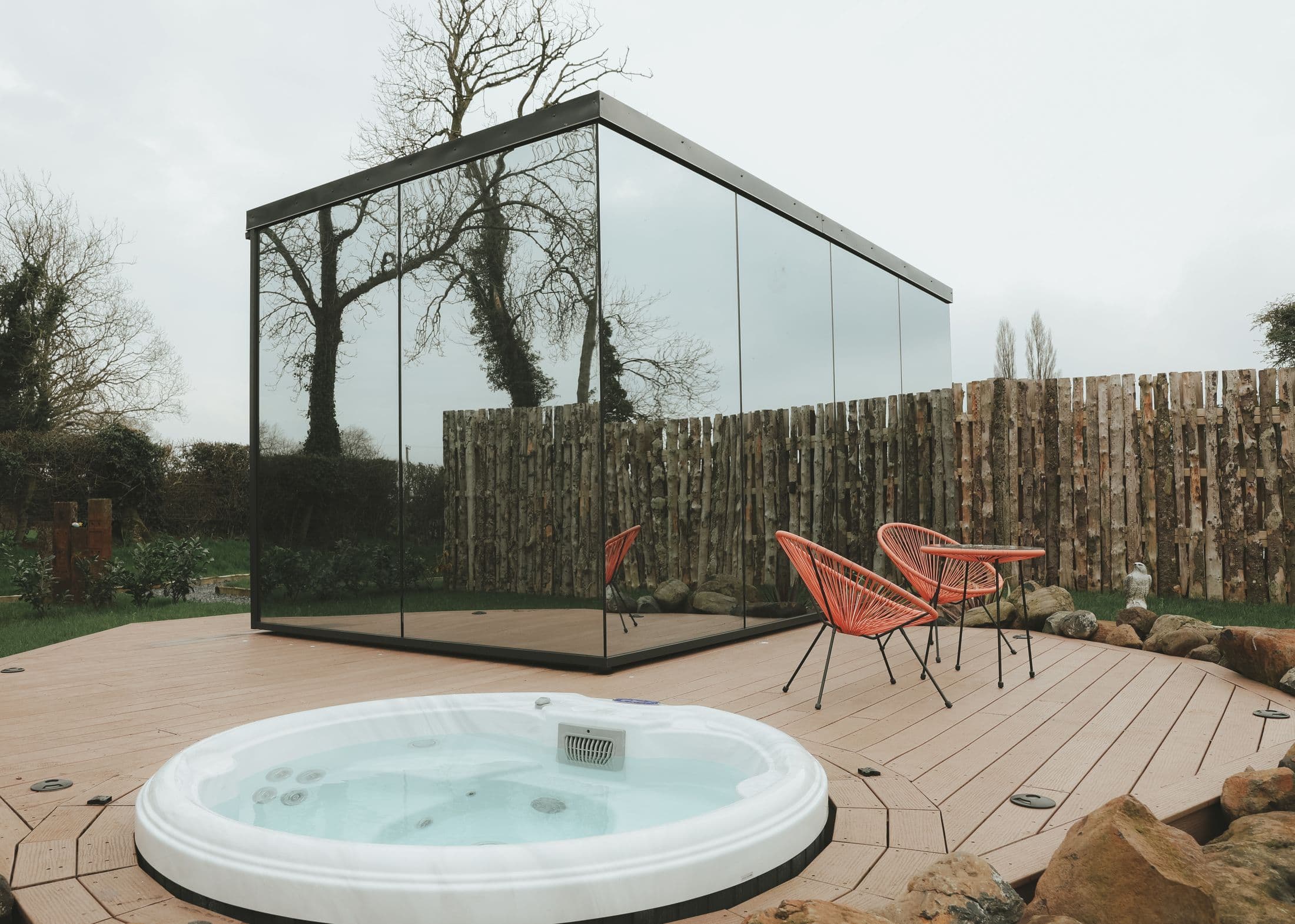 The Sunset mirror house reflecting the split-log fence and bare trees, with the round hot tub sunk into the timber decking in front